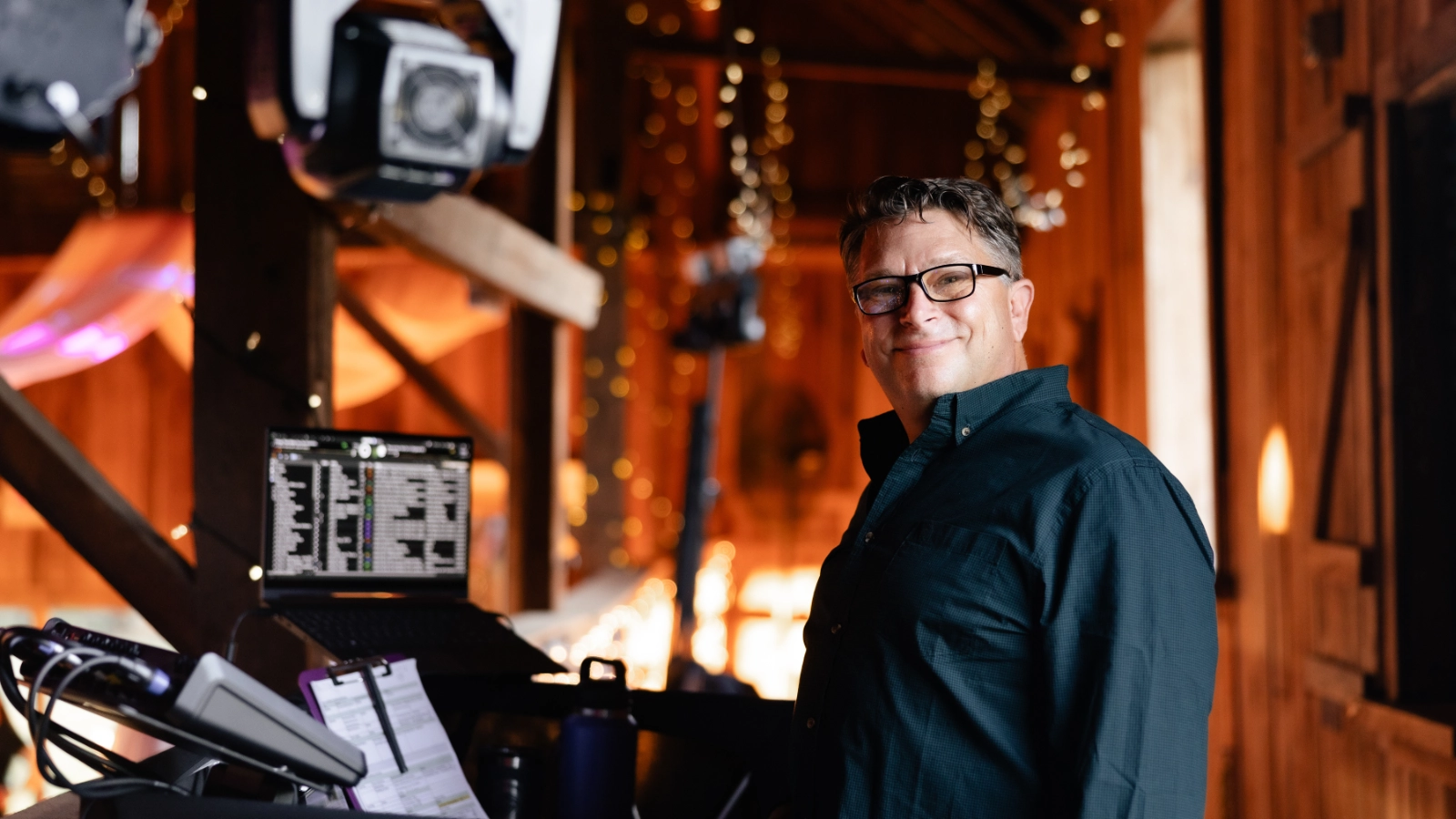 DJ Sideout standing beside the production booth at a warmly lit barn wedding.