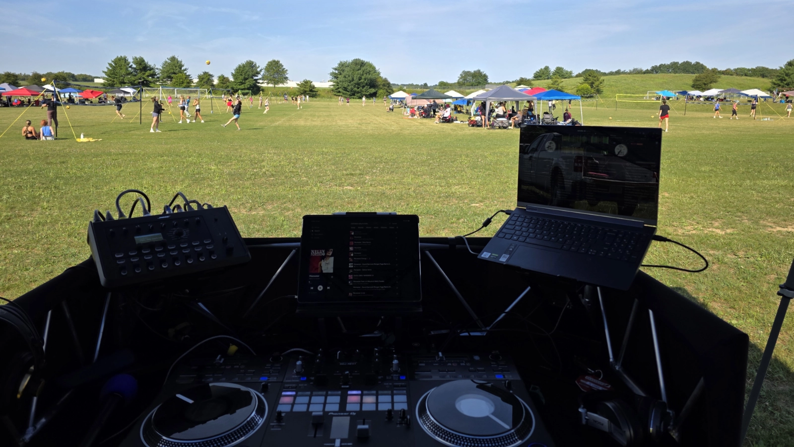 DJ production setup overlooking multiple outdoor volleyball courts, team tents, and tournament activity.