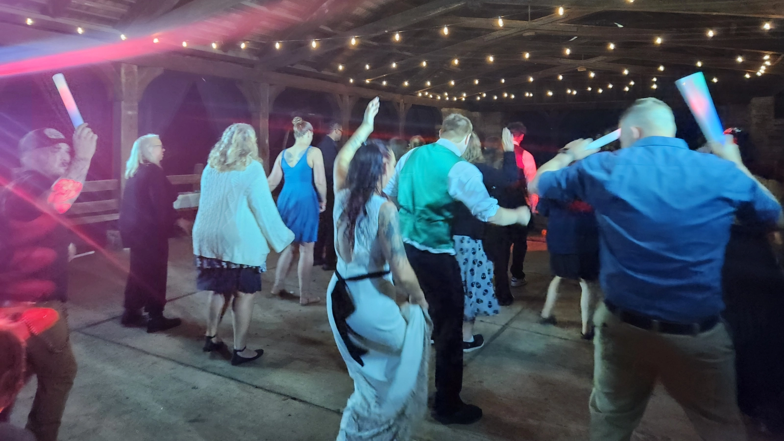 Wedding guests dancing under string lights and colorful event lighting at an open-air reception.