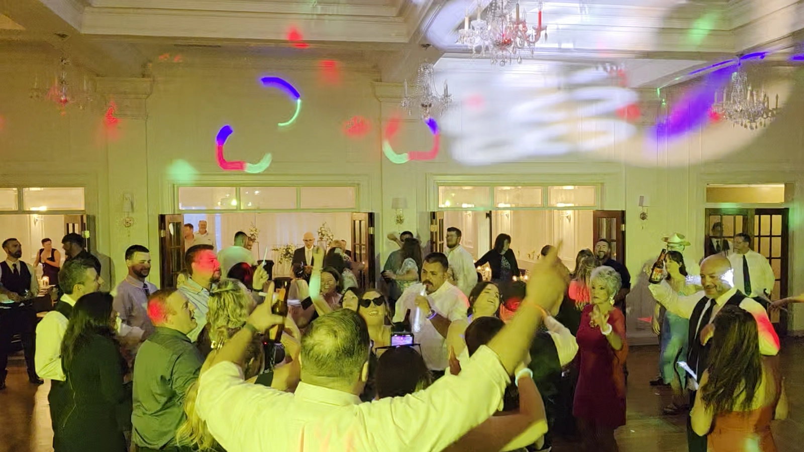 DJ Sideout wedding dance floor with guests celebrating beneath colorful moving-light effects in an elegant ballroom.