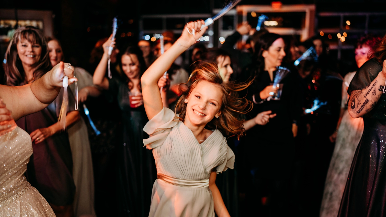 A young wedding guest dancing enthusiastically with a light wand as other guests celebrate around her.
