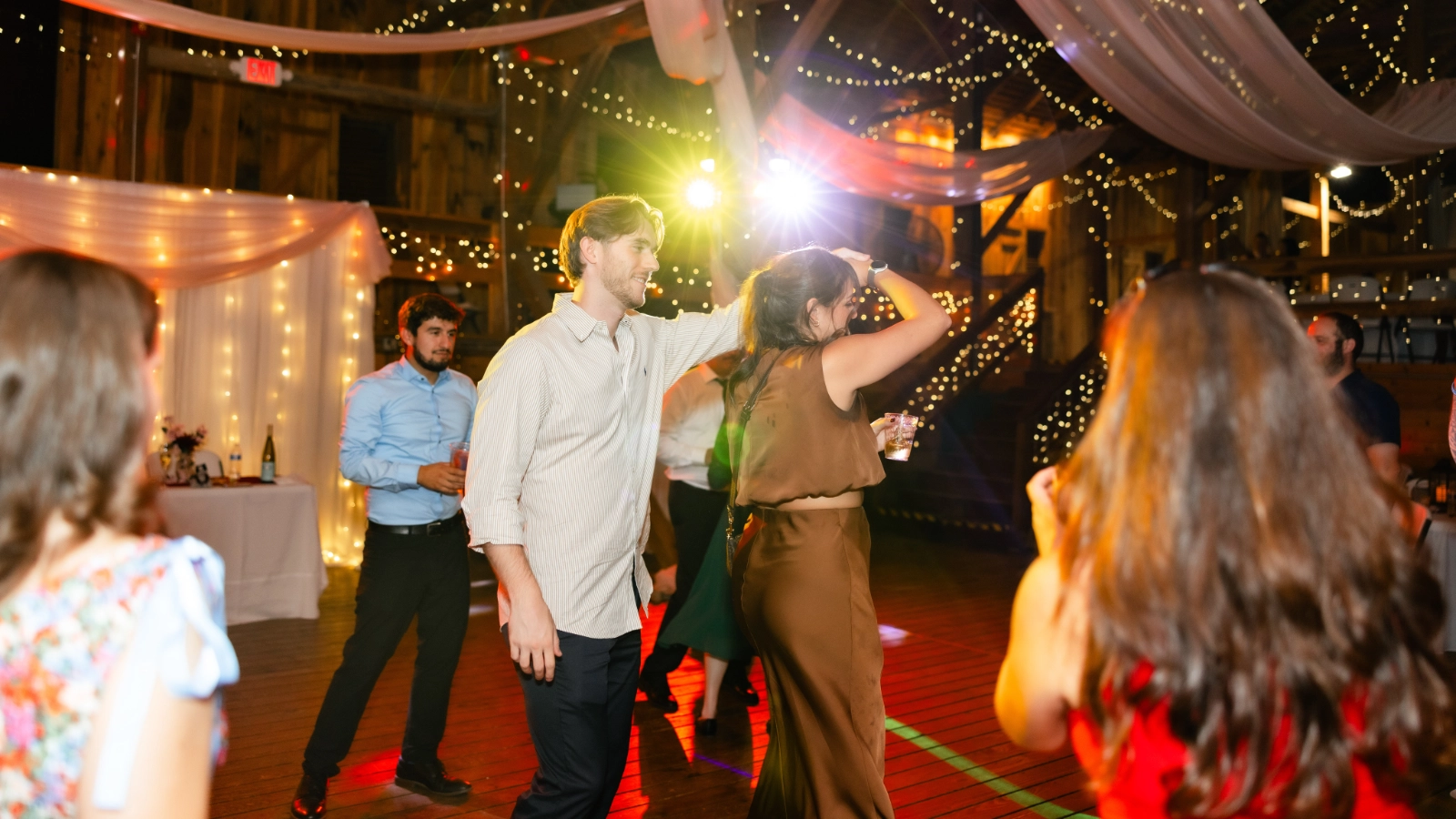 Wedding guests dancing beneath warm string lights and colorful DJ lighting in a rustic barn reception.