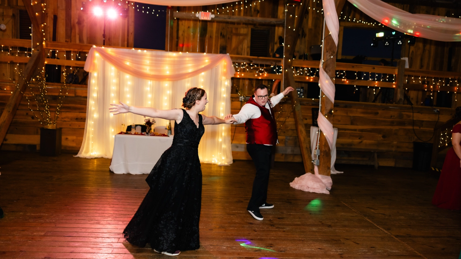 A newlywed couple dancing together beneath warm string lights in a rustic barn reception.